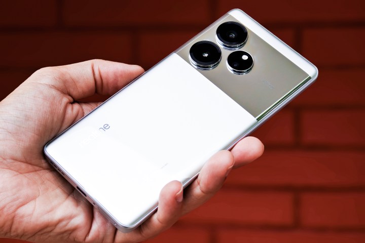 Realme GT 6 Android phone in silver color held in hand in front of a red brick wall.