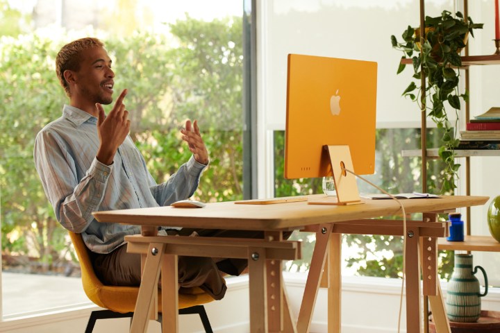 A man sitting at a desk in front of an M1 iMac. Behind him is a large glass window and a set of shelves holding books, plants and ornaments.