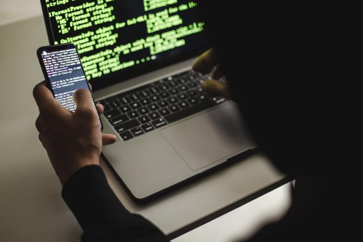 A hacker typing on an Apple MacBook laptop while holding a phone. Both devices show code on their screens.