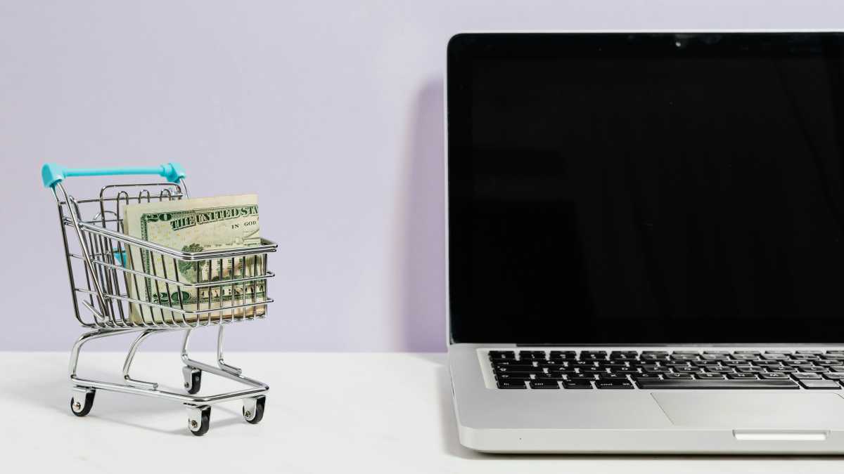 Alt: A laptop with a blank screen sits next to a miniature shopping cart filled with cash, illustrating the potential for selling used laptops.