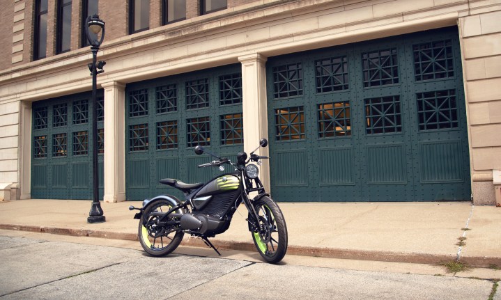 Alt: A Royal Enfield Flying Flea FFC6 electric motorcycle parked on the curb in front of a garage.