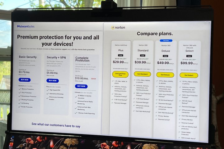 A PC monitor shows a side-by-side comparison of Malwarebytes and Norton pricing.