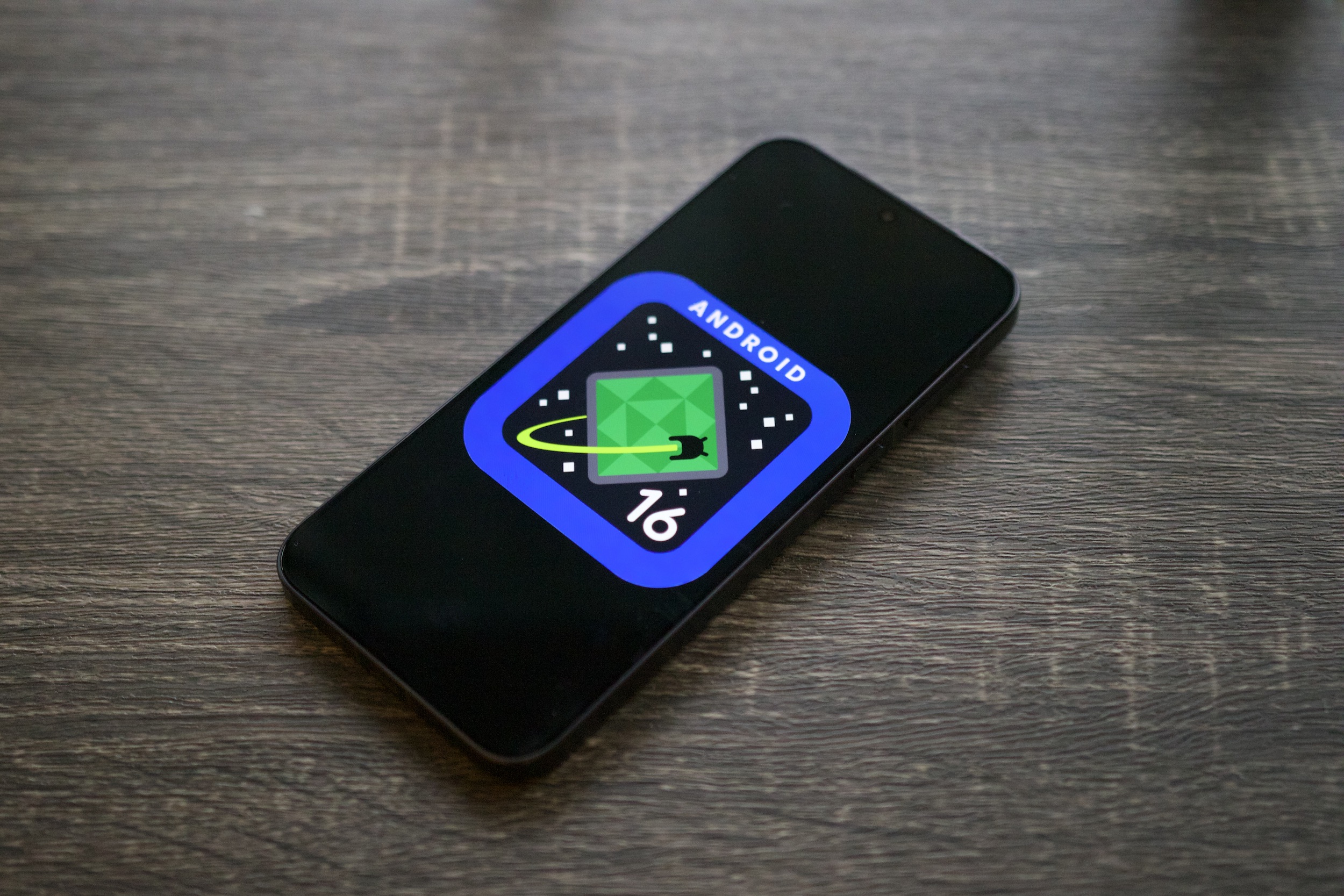 The Android 16 logo on a smartphone, resting on a shelf.